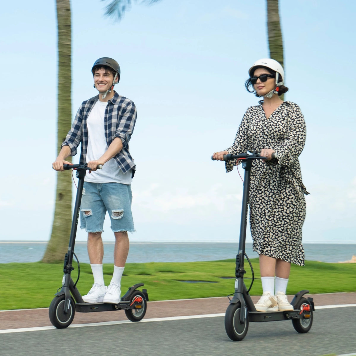 Two riders on black electric scooters with 10-inch tires and rear suspension on a seaside path.