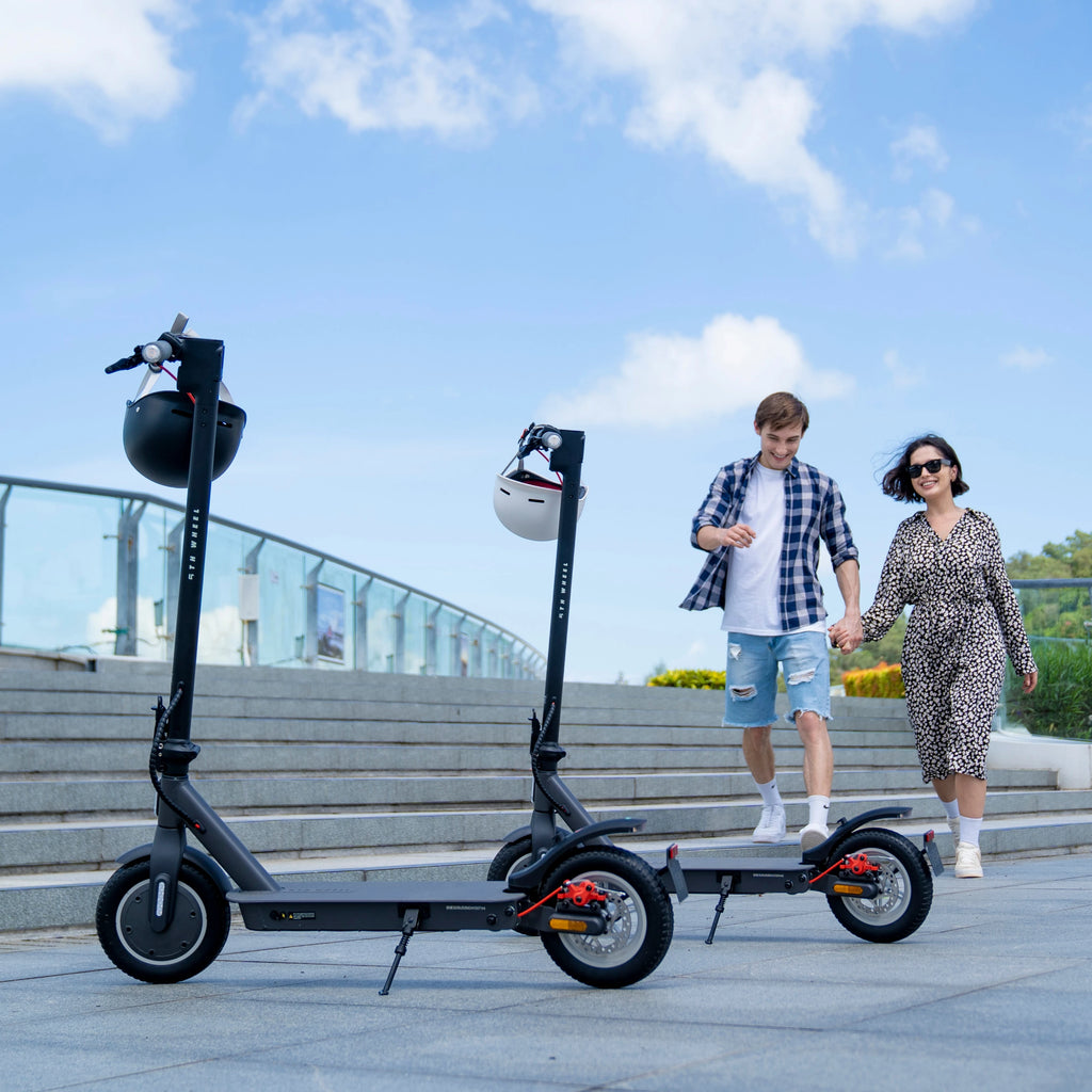 Two black electric scooters with 10" tires and helmets on handlebars, parked near outdoor stairs with a couple walking.