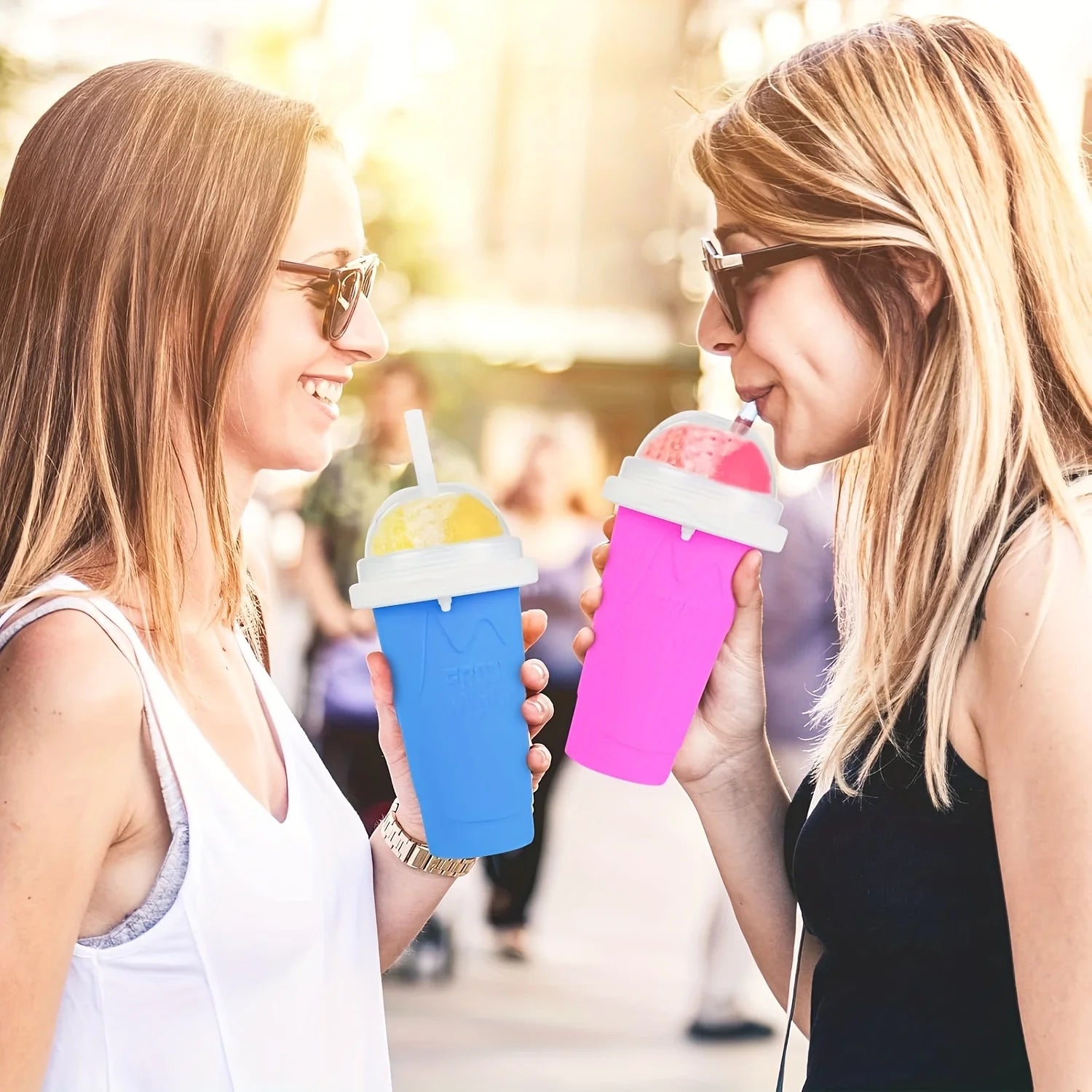 Two women holding a blue and a pink squeeze cup with slushy drinks, complete with white lids and straws.