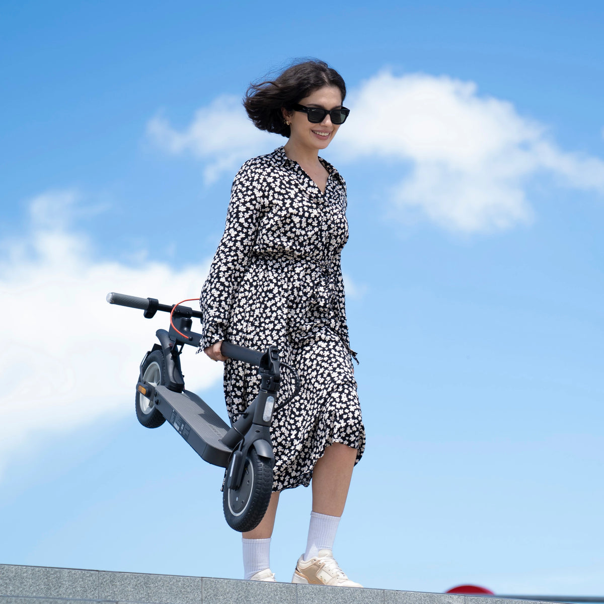 A smiling woman carries a dark grey 5TH WHEEL V30Pro electric scooter with large tires outdoors.