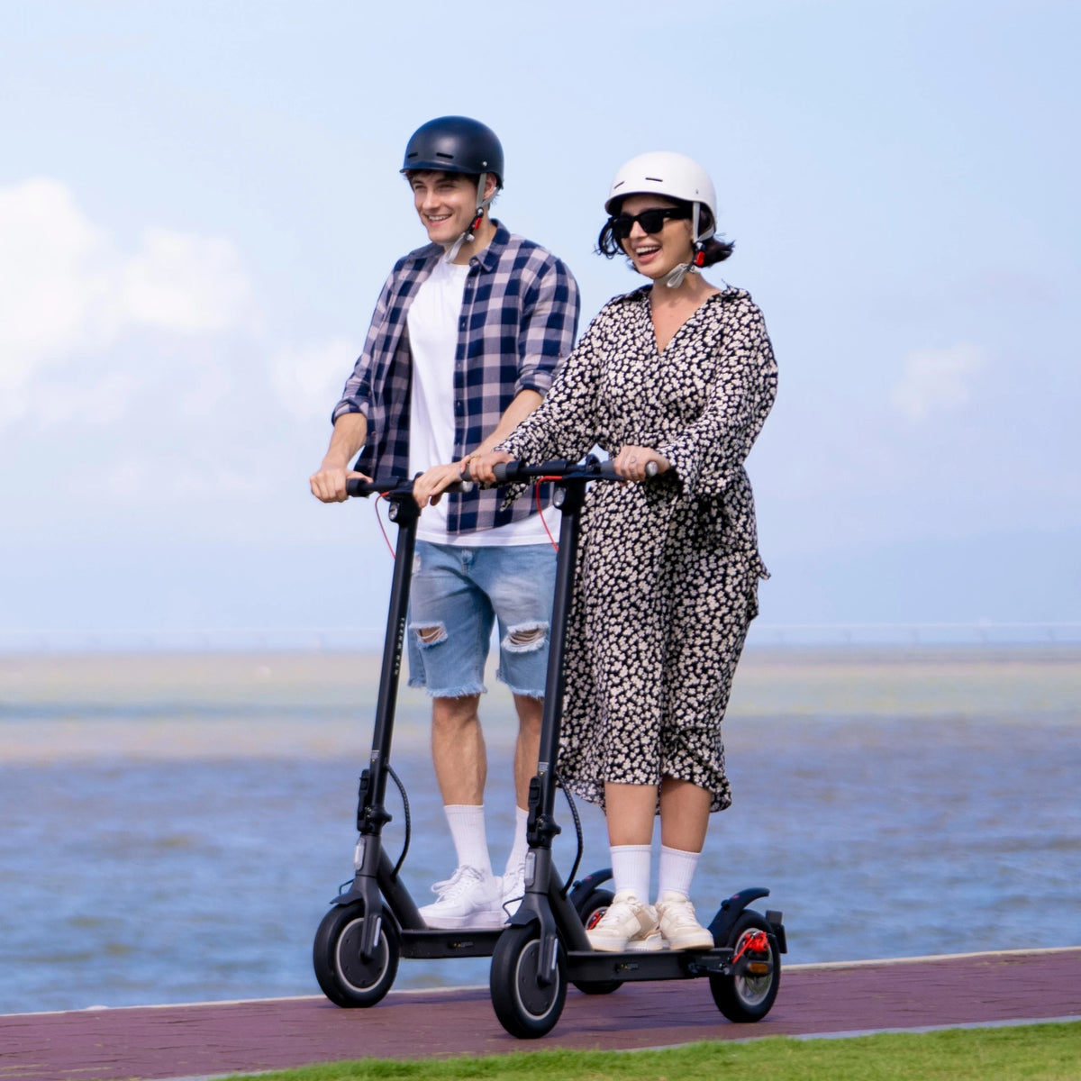 Two people wearing helmets ride black 5TH WHEEL V30Pro electric scooters on a paved path near the water.