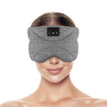A woman wearing a grey, padded wireless sleep mask with built-in Bluetooth headphone controls on the front.