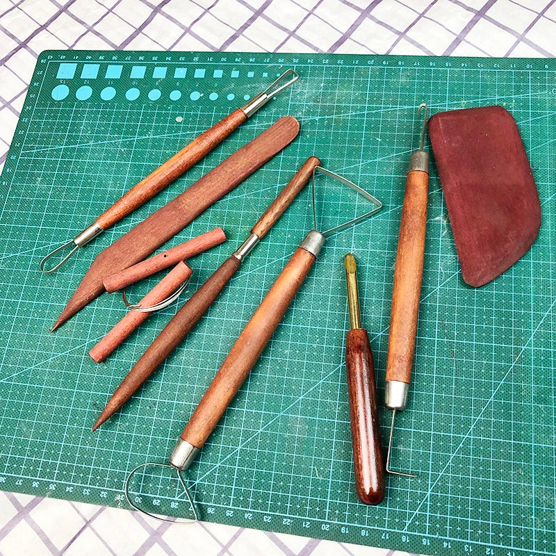 Set of wooden-handled pottery sculpting tools including wire loops, carving blades, and a rubber rib on a cutting mat.