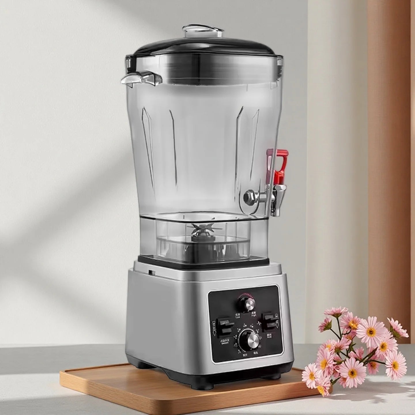 Silver commercial blender with a clear pitcher, red dispensing tap, and manual control knobs on a wooden tray.