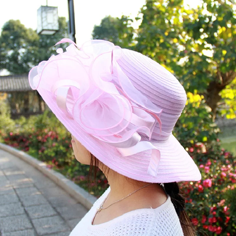 A pale pink wide-brim occasion hat featuring elegant layered organza flower detailing.