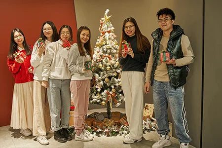 A group of people standing around a decorated Christmas tree, holding small wrapped gifts.