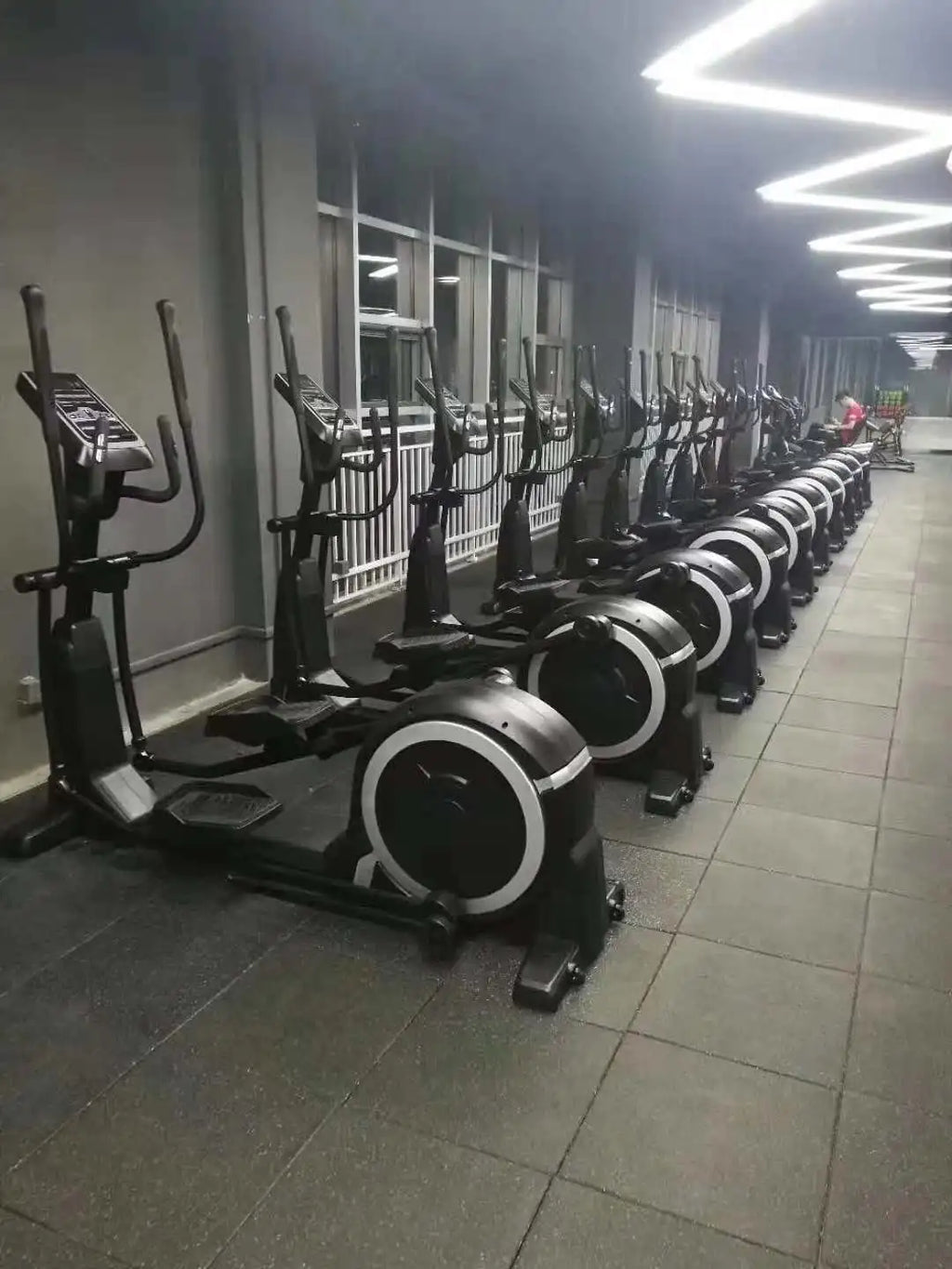 A row of black commercial indoor elliptical trainers with circular white-rimmed flywheels in a modern gym setting.