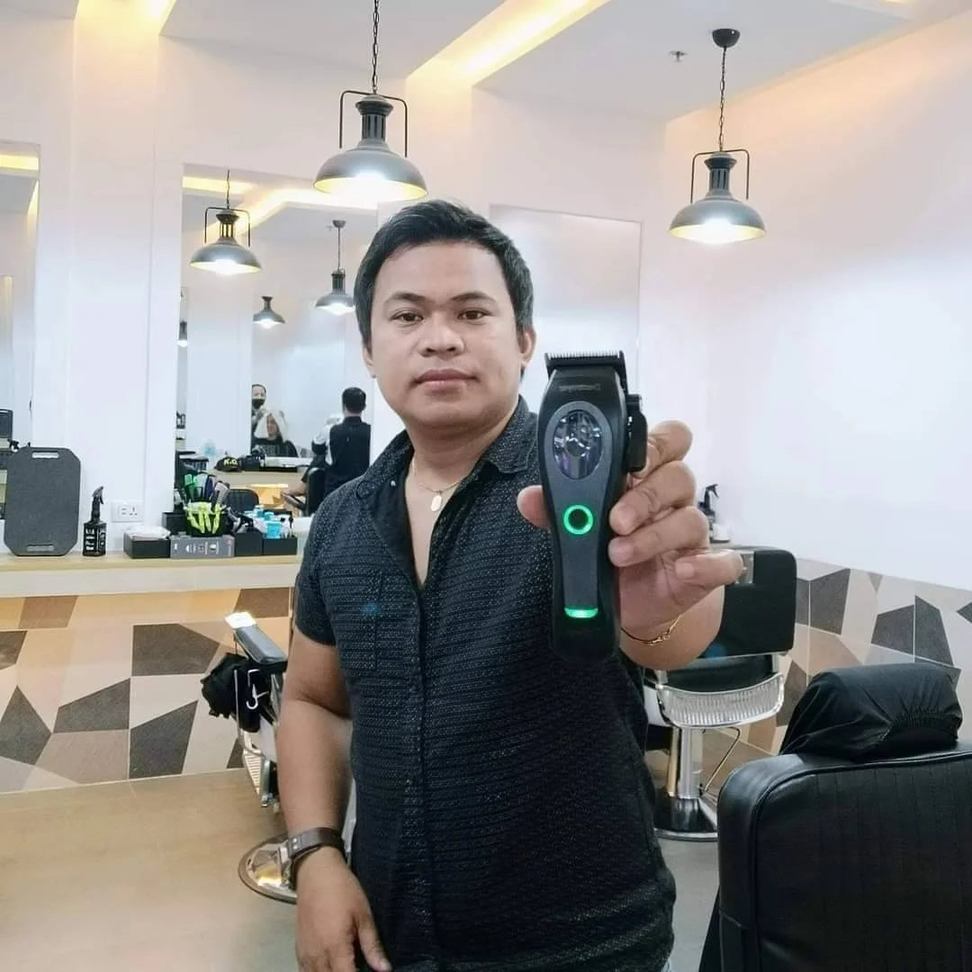 A barber holding a matte black, cordless hair clipper featuring a circular green LED power indicator light.