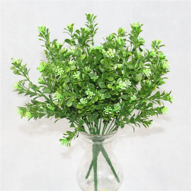 A bouquet of artificial green plastic plants with small, light-colored buds in a clear glass vase against a white wall.