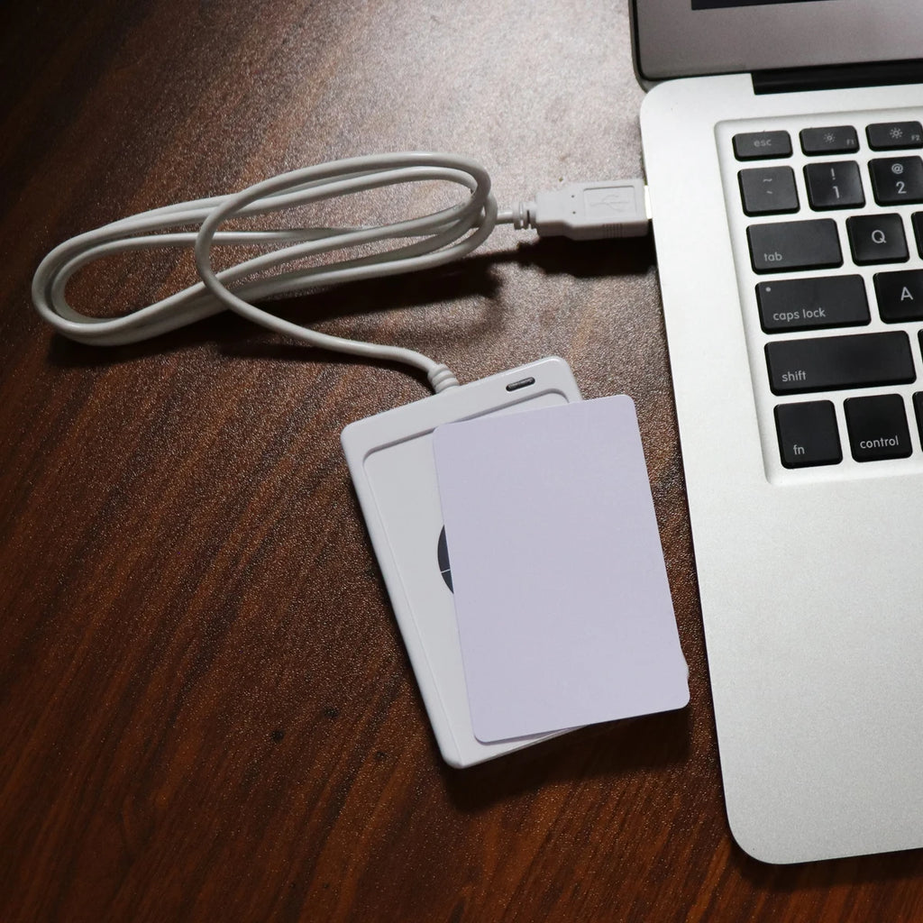 White plastic RFID card reader connected via USB cable to a silver laptop on a wooden surface with a blank white card.