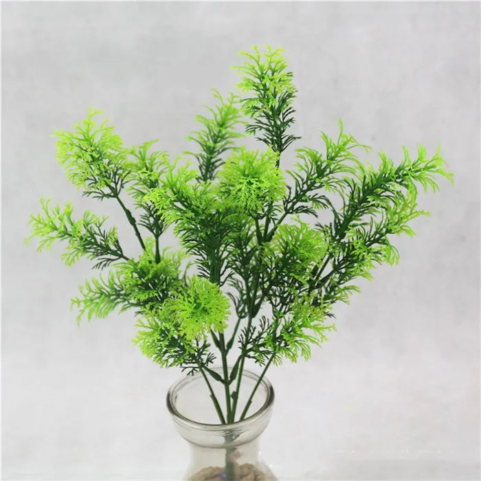 A bright green artificial fern sprig with wispy, needle-like foliage, displayed in a clear glass vase.