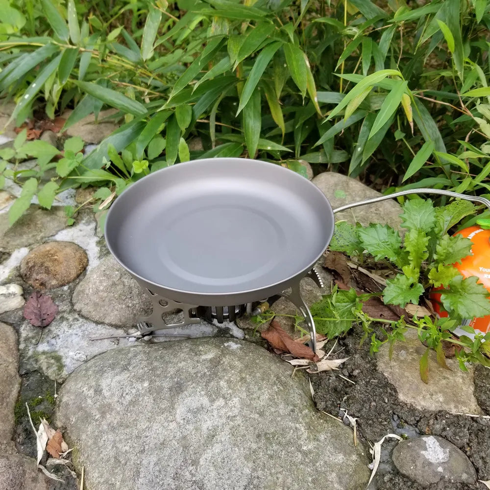 A grey titanium frying pan sits on a portable camping stove over rocks and green foliage.
