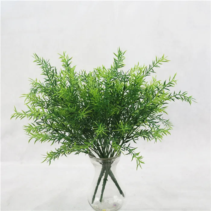 A bouquet of lush green artificial plastic fern grass in a clear glass vase against a plain white background.