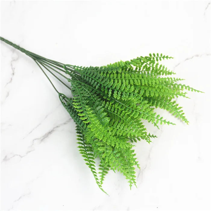 Artificial bright green fern fronds with delicate, serrated leaves on long stems against a white marble background.