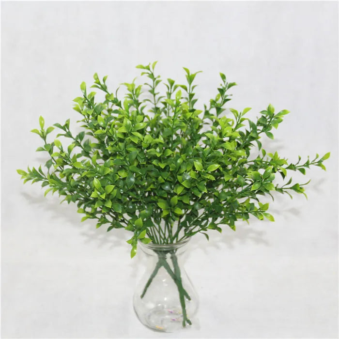A bunch of vibrant, artificial green leafy branches arranged in a clear glass vase against a white background.