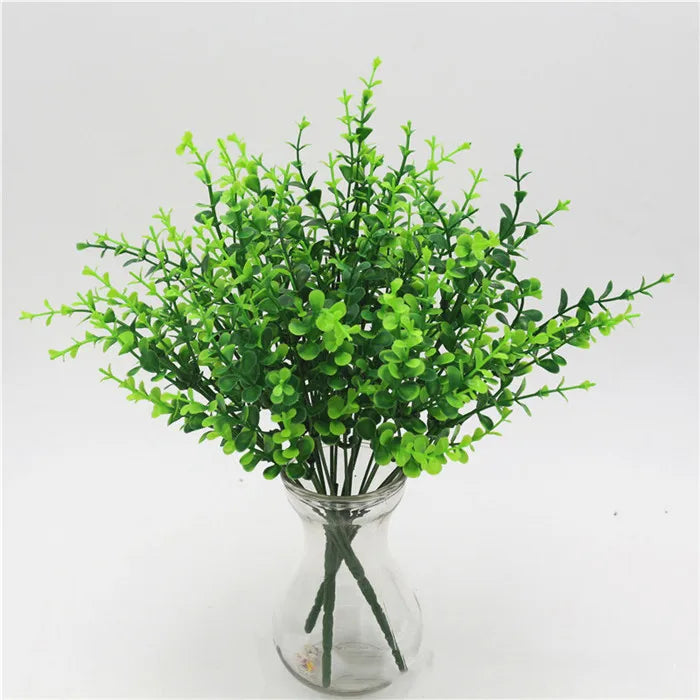 A bunch of vibrant green artificial leafy stems in a clear glass vase against a white background.