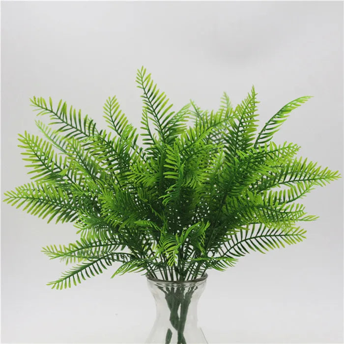 A vibrant green artificial fern bush with feathery, pointed leaves in a clear glass vase against a white background.