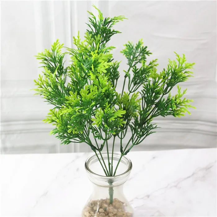 Artificial green fern sprig with feathery leaves in a glass vase on a white marble surface.