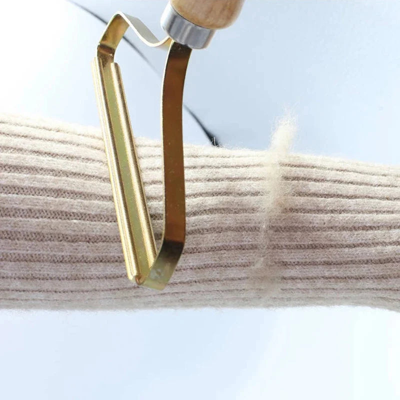 Gold metal fabric lint remover with a wooden handle, being used to remove bobbles from a cream ribbed jumper sleeve.