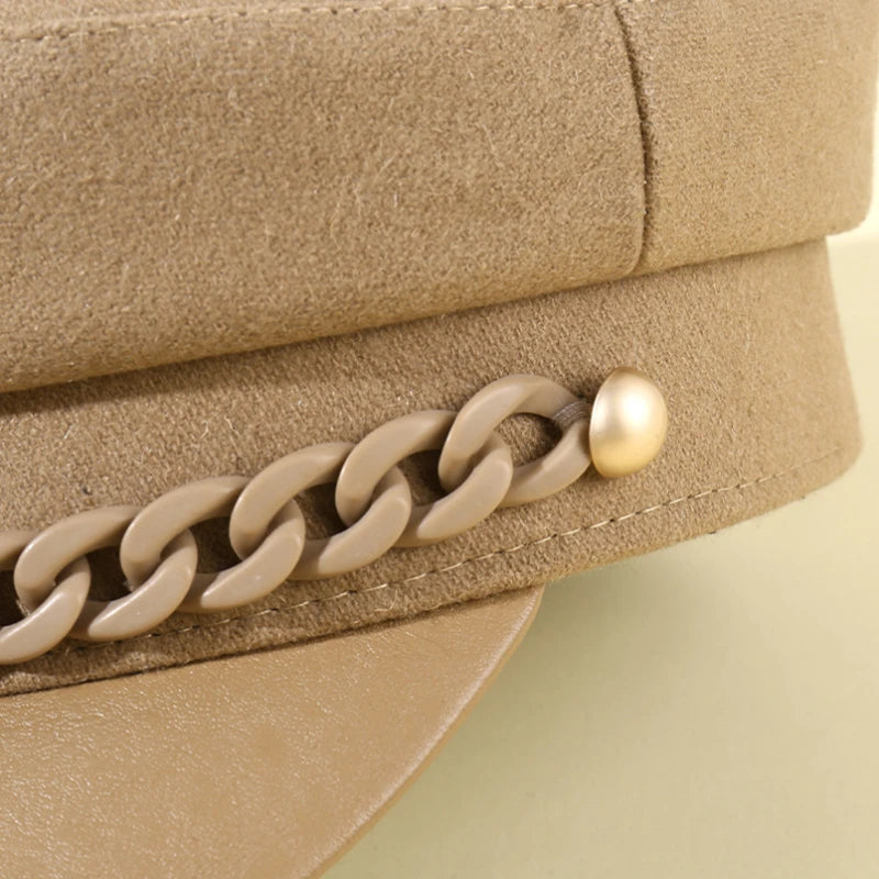 A close-up of a camel-coloured felt baker boy hat, featuring a decorative matching chain and gold-tone stud detail.