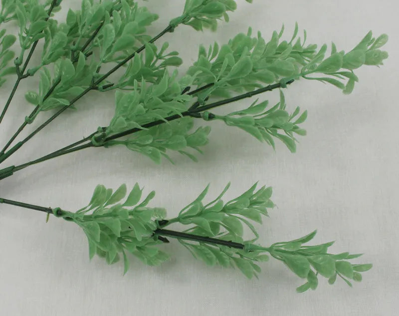 A sprig of artificial light green plastic fern-like leaves on dark stems against a plain off-white background.