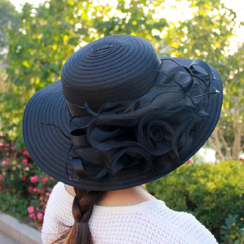 A woman wearing a wide-brimmed black occasion hat featuring an elegant, layered organza floral side embellishment.