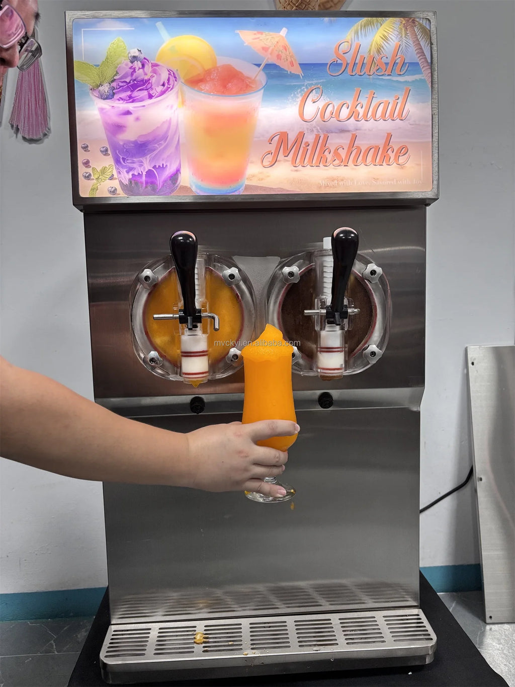 Silver commercial two-tank slushy machine with black dispensing handles, pouring orange slush into a glass.