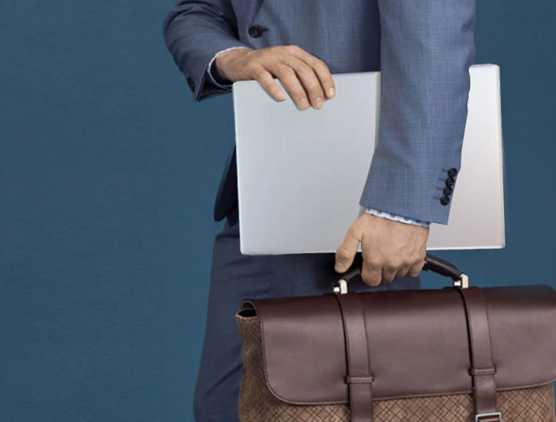 A professional in a blue suit holding a sleek silver 15.6-inch laptop and a brown leather briefcase.