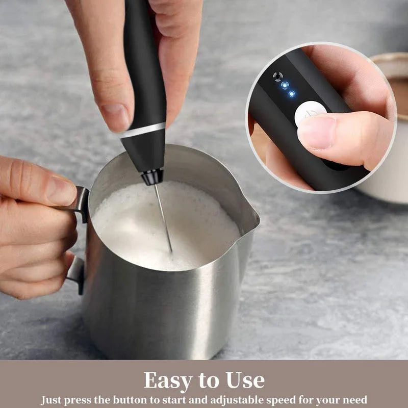 Black handheld milk frother with a silver metal whisk, shown frothing milk in a stainless steel pitcher.