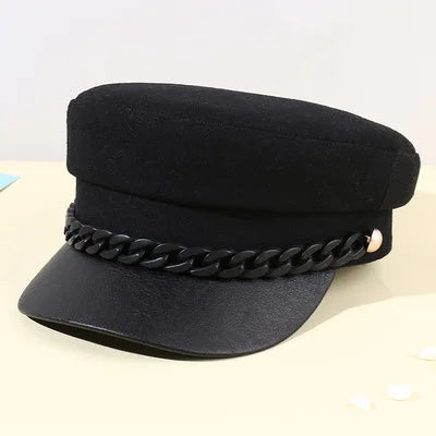 Black baker boy cap with a flat top, stiff peak, and a decorative chunky black chain detail across the front.