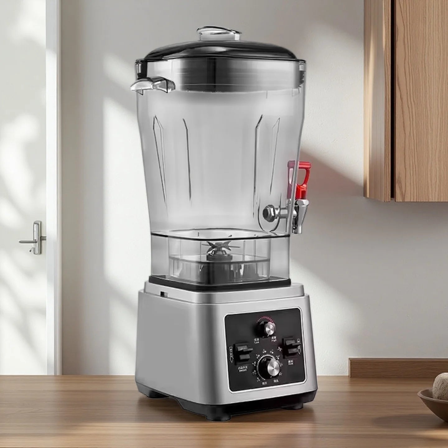 Silver commercial blender with a large clear pitcher, dispensing tap, and manual dial controls on a wooden countertop.