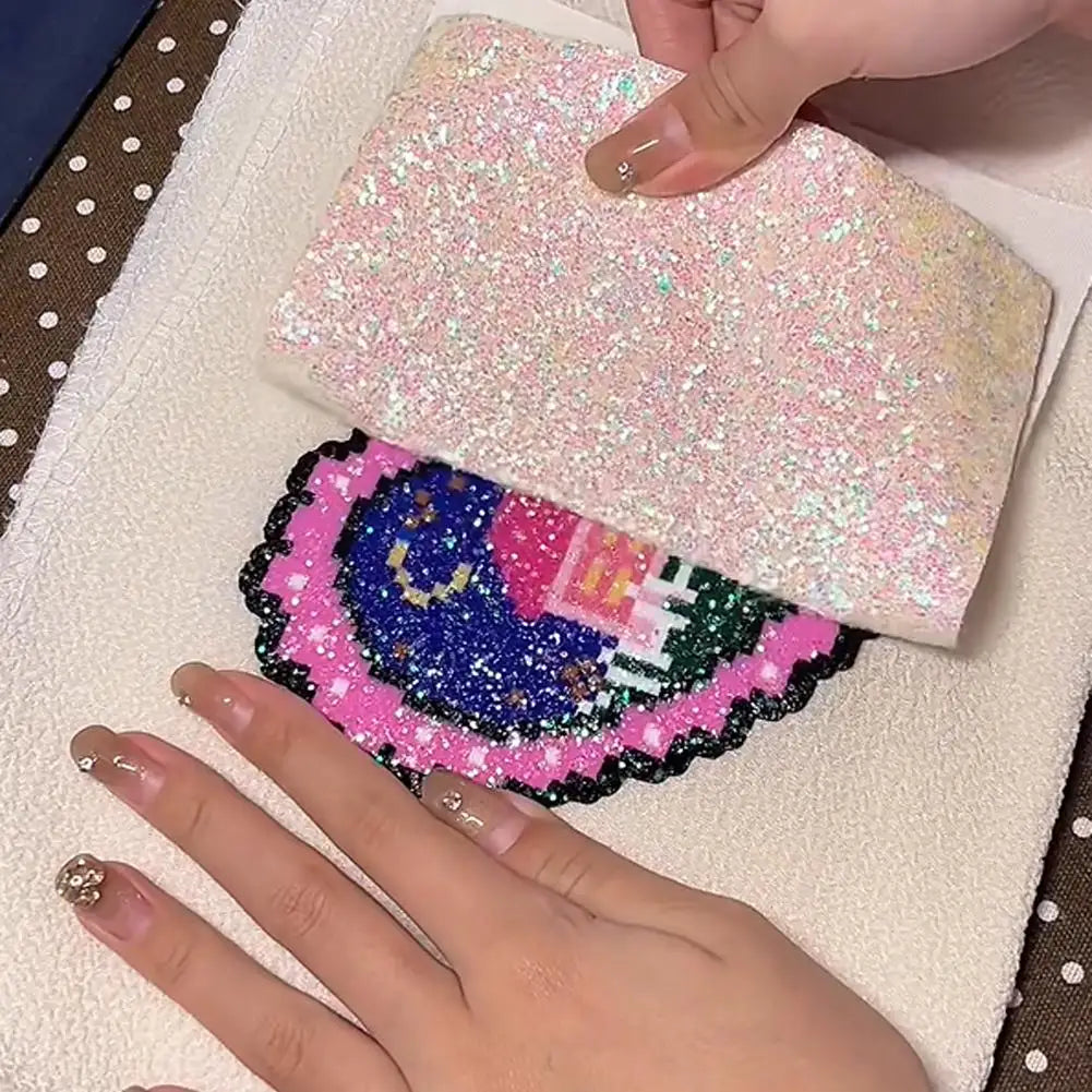 Hands lifting a sheet of iridescent glitter fabric to reveal a colourful, sparkling pixel-art embroidery underneath.