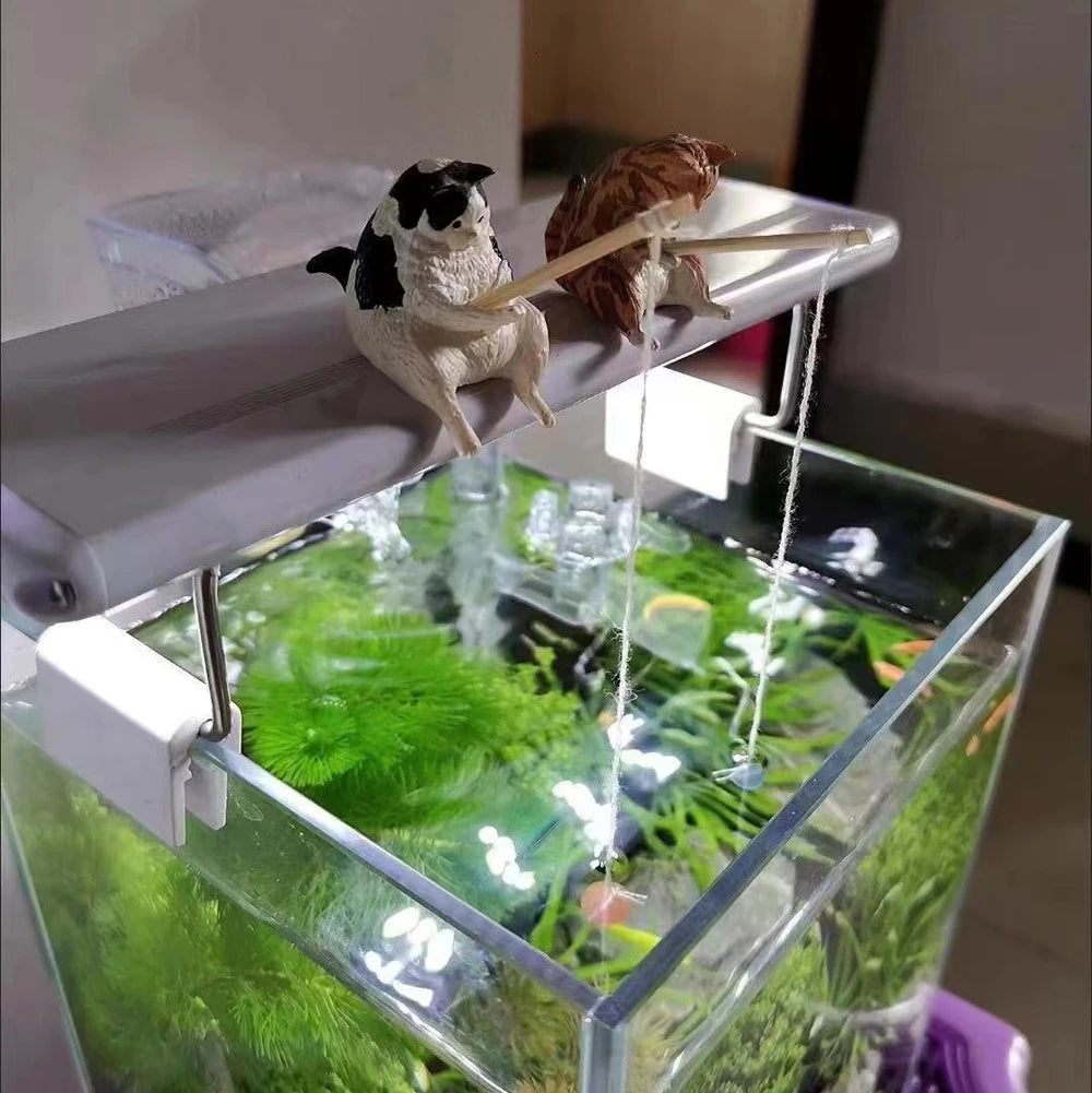 Two small, detailed cat ornaments, one black-and-white and one ginger, sitting on an aquarium light, fishing.