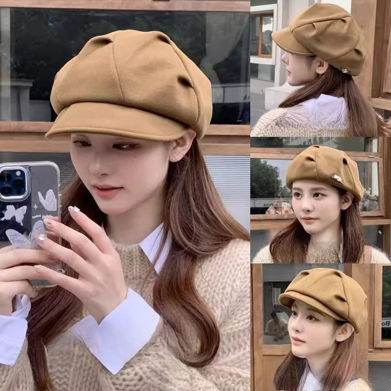 A woman wearing a tan, structured newsboy beret with a curved peak and soft, pleated crown detailing.