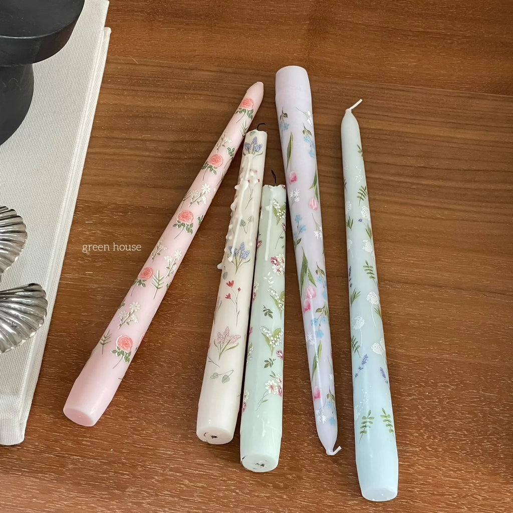 Five tapered dinner candles in pastel colours with delicate floral patterns on a wooden surface.