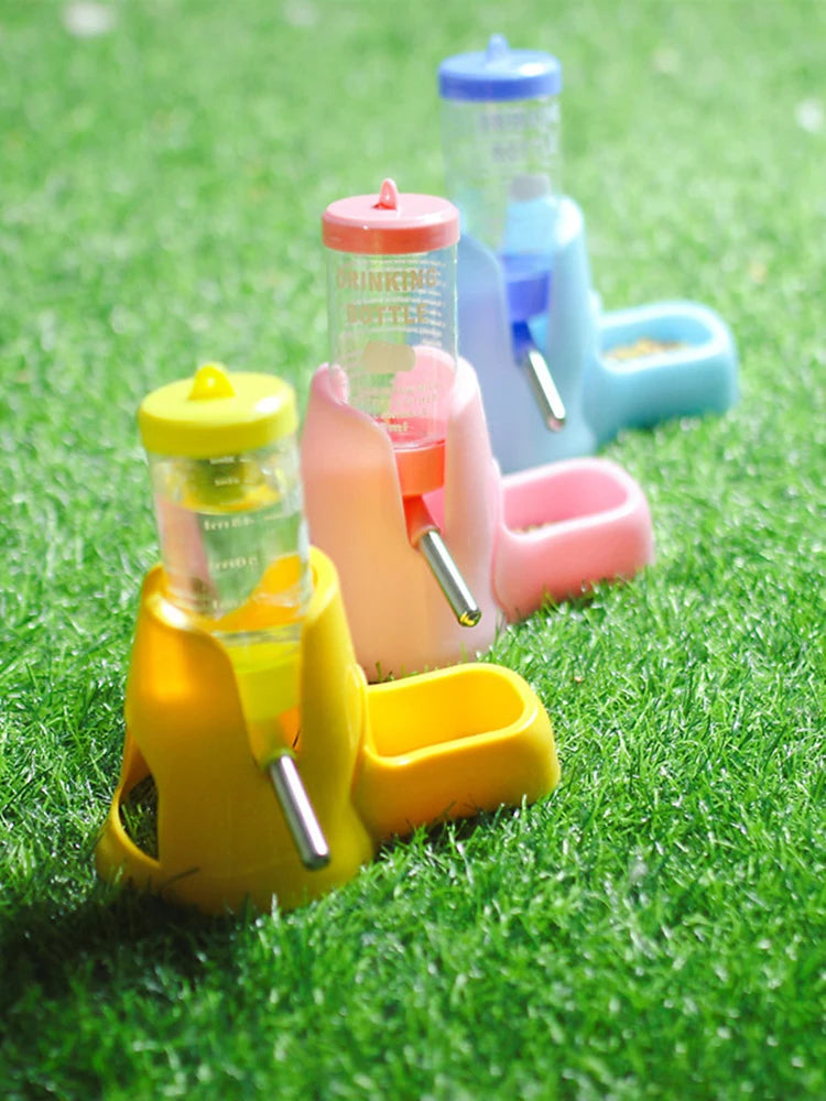 Three colorful, plastic small pet water dispensers with built-in food bowls, set on green artificial grass.