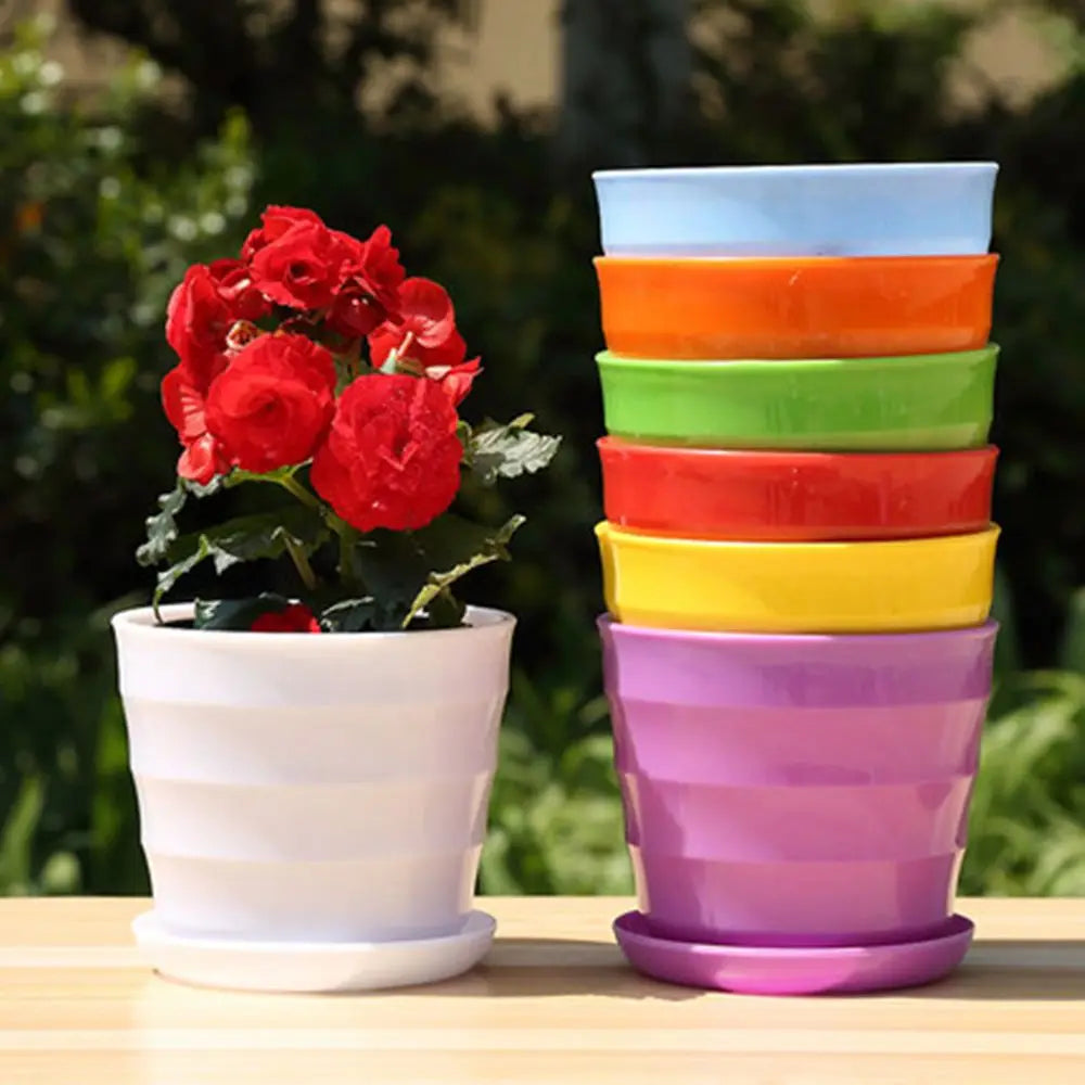 A red flowering plant in a white pot beside a stacked set of colorful plastic plant pots with matching saucers.