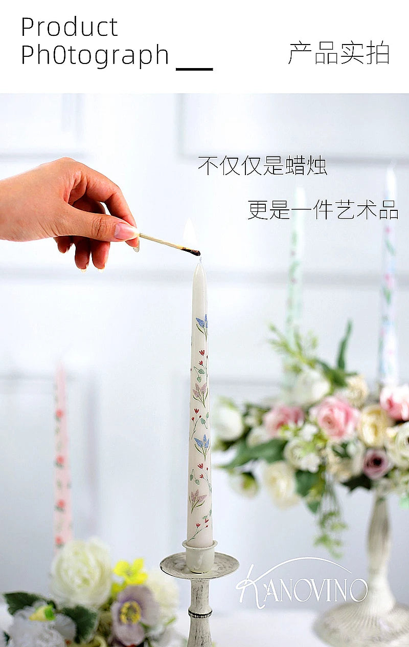 A hand lights a tall, cream-coloured tapered candle featuring a delicate floral pattern, set in an antique holder.