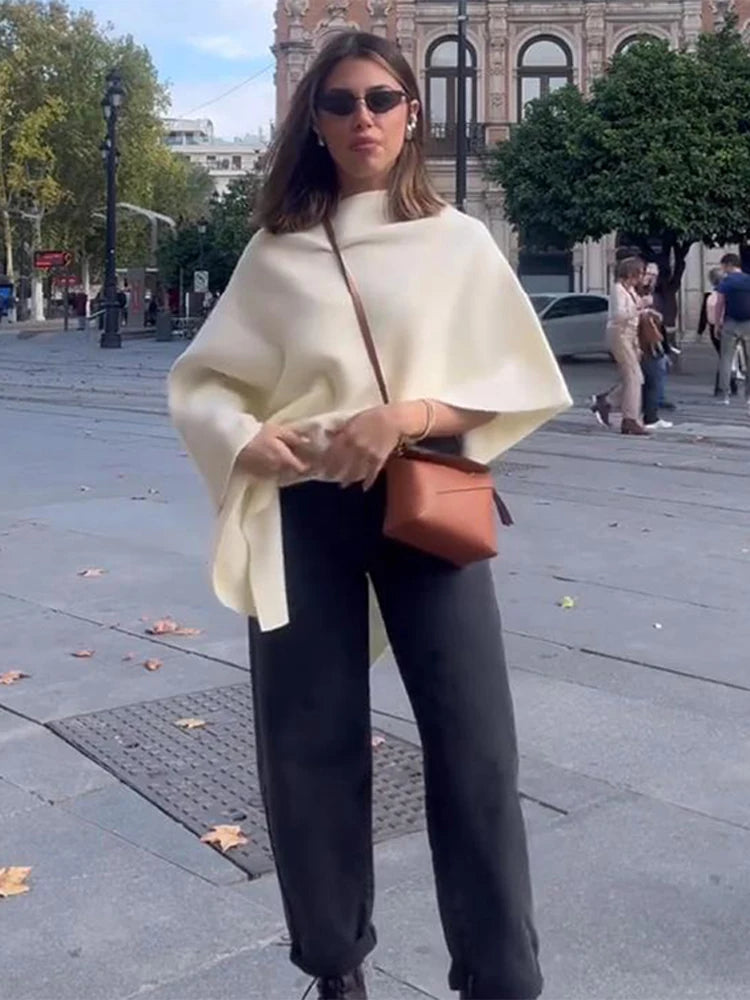 Woman wearing a loose cream knitted poncho style sweater with irregular draping and wide sleeves.