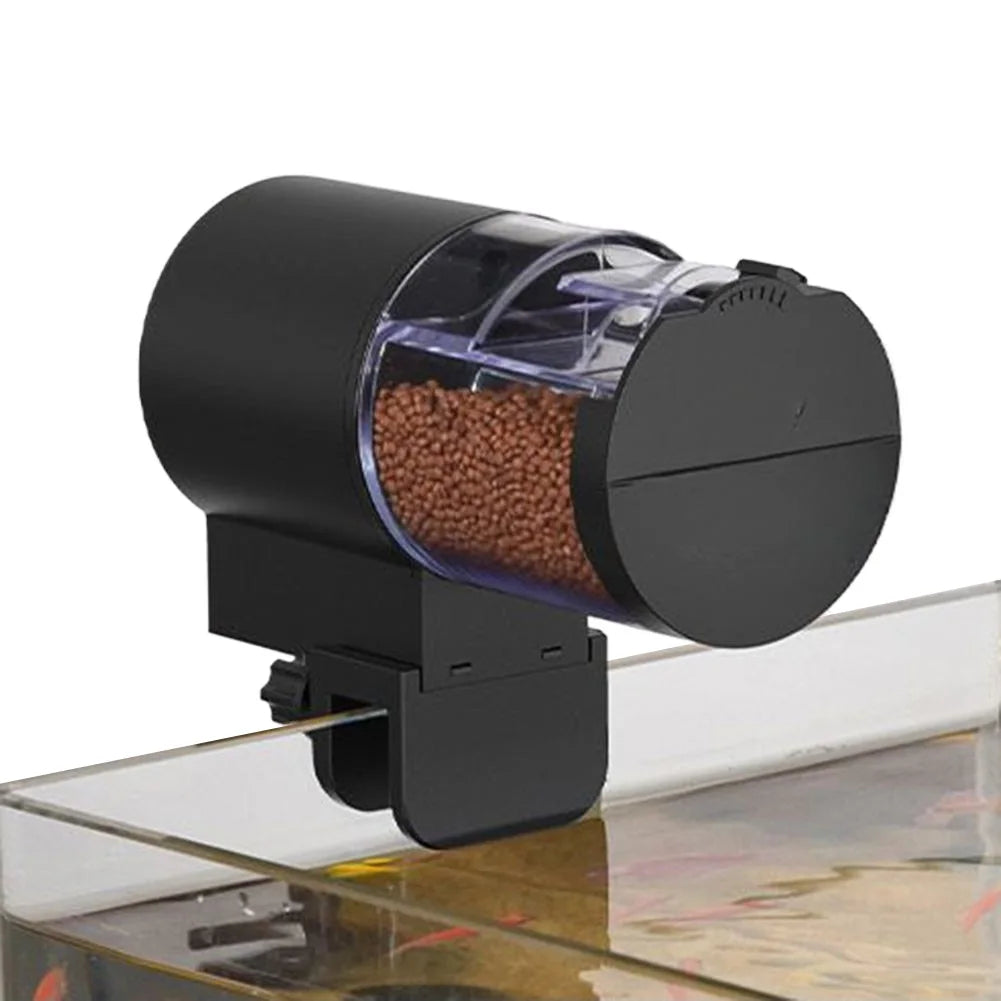 Black automatic fish feeder with a transparent, cylindrical food container mounted on the edge of a glass aquarium.