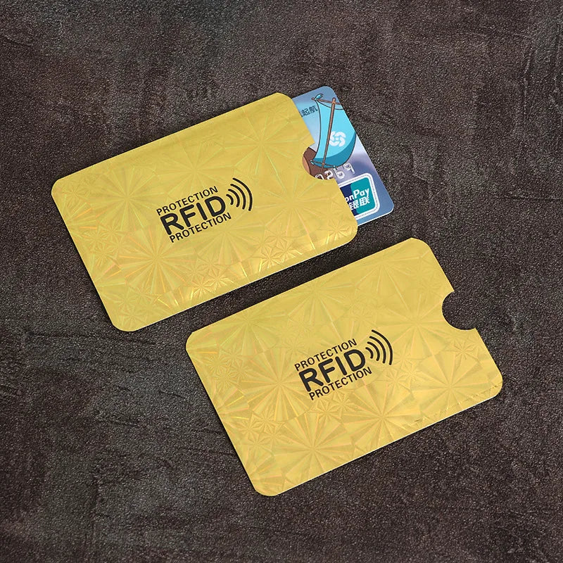 Two gold, metallic RFID-blocking card sleeves with geometric patterns and thumb notches for sliding cards out.