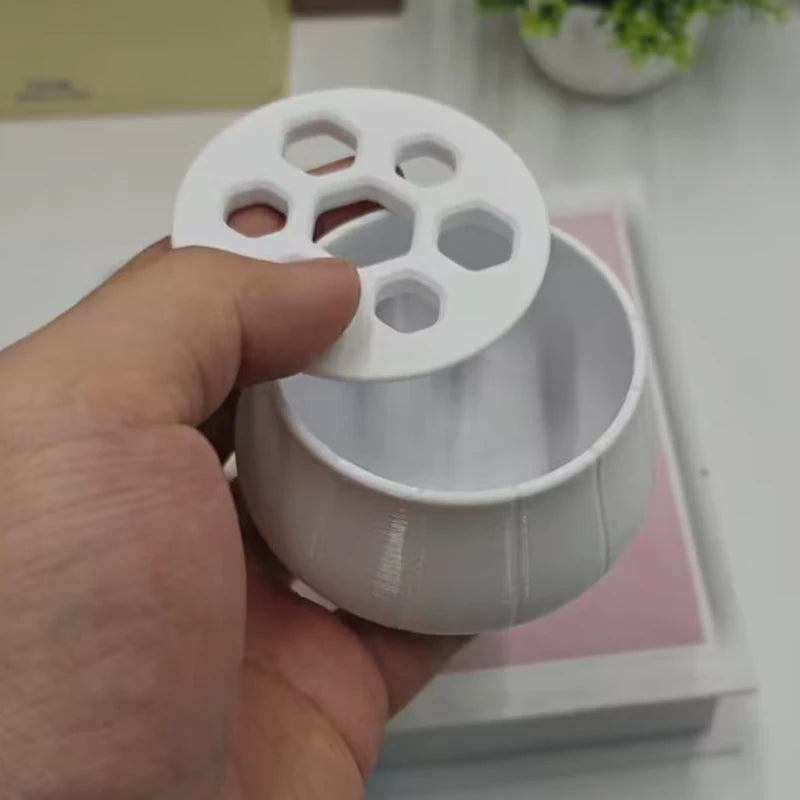 A hand holds a white, 3D-printed hydroponic planter with a circular lid featuring hexagonal cutouts for plant stems.