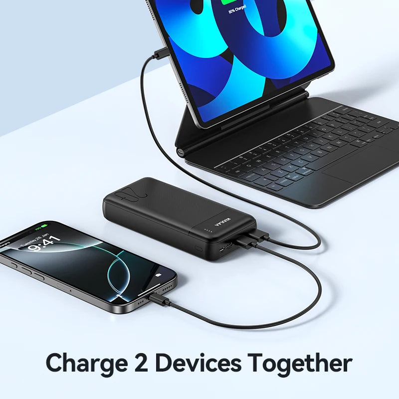 A black, rectangular 20,000mAh portable power bank with an LED indicator charging a smartphone and tablet simultaneously.
