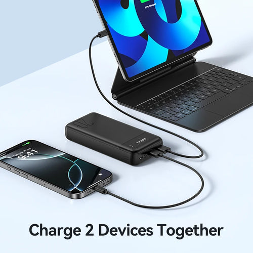 A black, rectangular 20,000mAh portable power bank with an LED indicator charging a smartphone and tablet simultaneously.