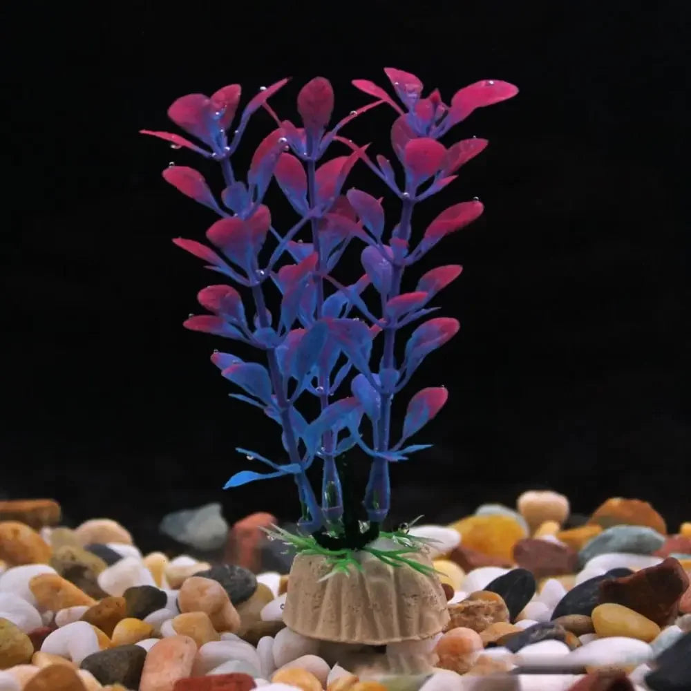 Artificial aquarium plant with blue and pink leaves on a weighted base, standing in colorful gravel on a black background.