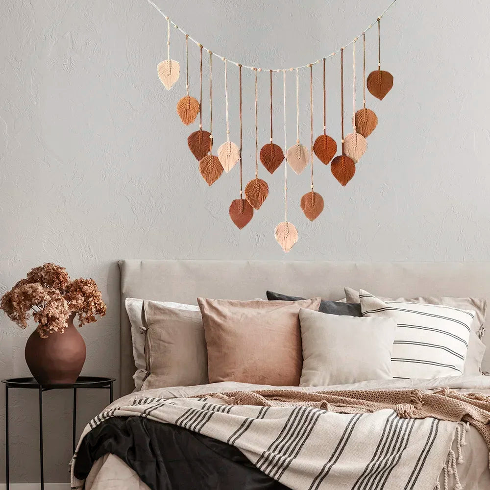 Boho wall hanging featuring macrame leaves in a cascading earth-tone gradient, suspended above a modern bedroom bed.