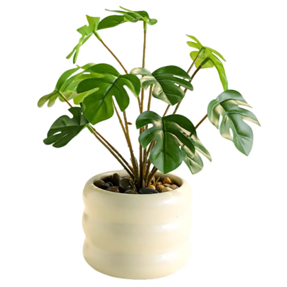 An artificial monstera plant with green, split leaves in a round, ridged cream ceramic pot filled with small stones.