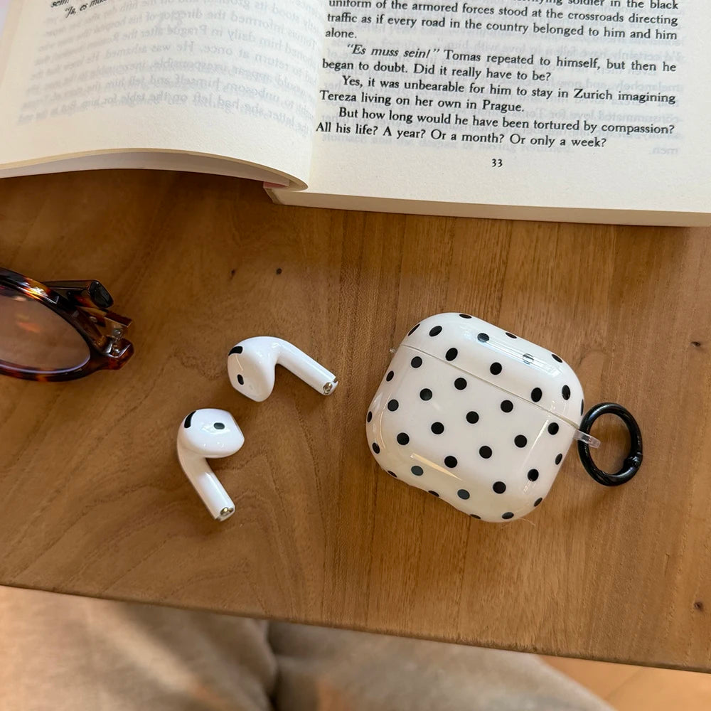 White polka-dot plastic case for AirPods, featuring a black carabiner clip, displayed on a wooden desk.