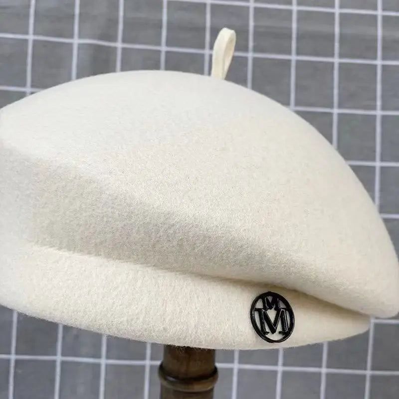 Cream wool-felt beret featuring a small top tab and a circular black 'M' logo detail on the side.
