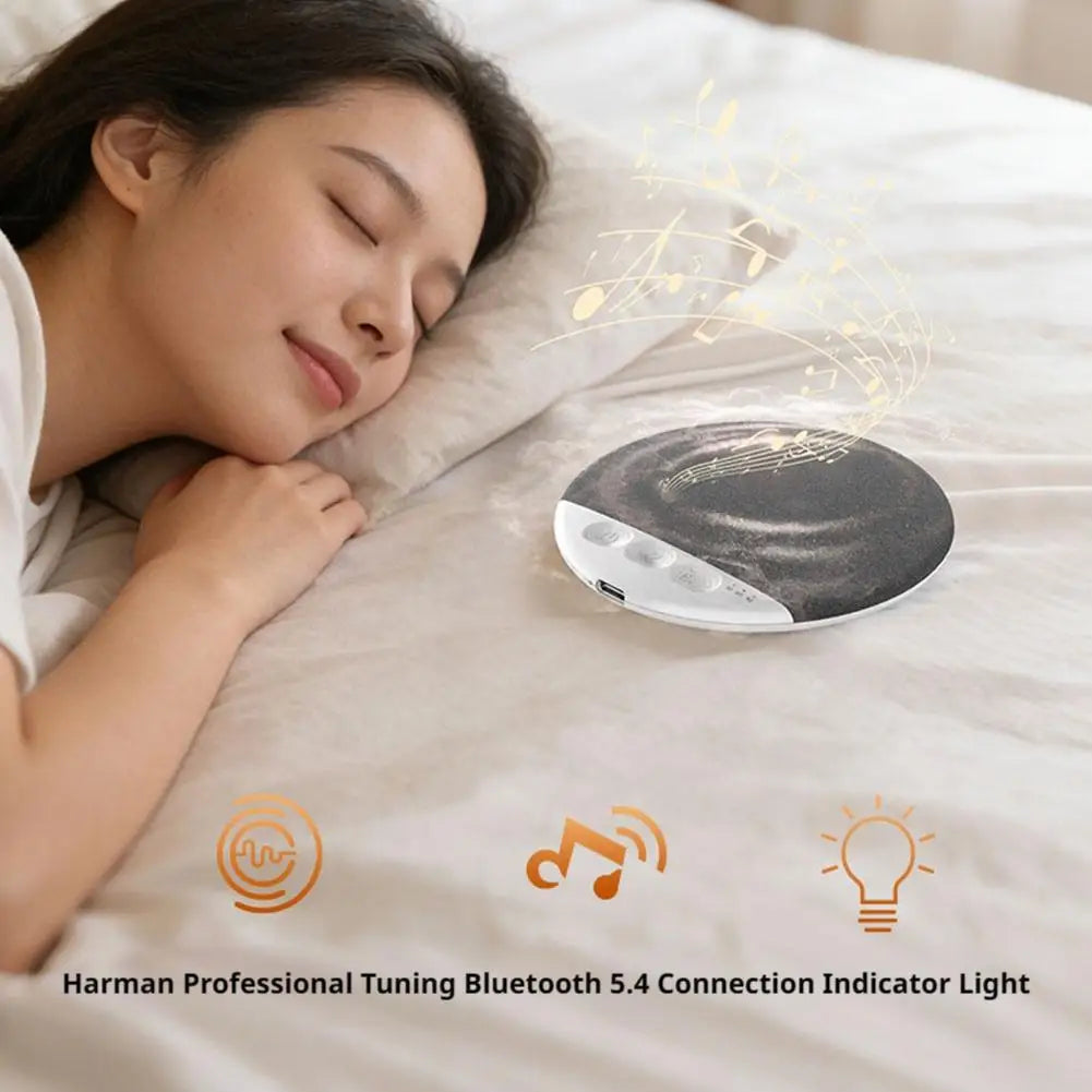 A person sleeping peacefully with a round, grey, wireless Bluetooth speaker resting on the white bedsheet.